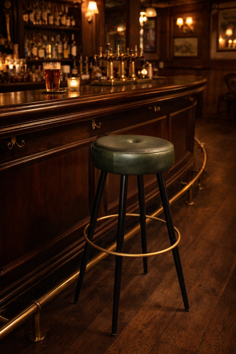 Taburete de bar de diseño vintage con asiento tapizado en cuero verde, patas negras y aro reposapiés dorado colocado en barra de pub escocés clásico.